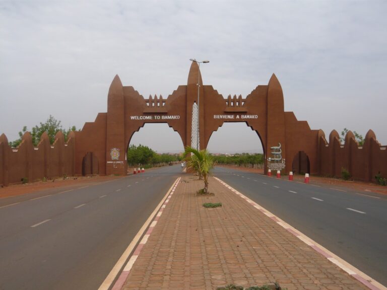 The image shows the entrance gate to Bamako, Mali, with "WELCOME TO BAMAKO" and "BIENVENUE A BAMAKO" inscribed on either side of the road leading into the city.