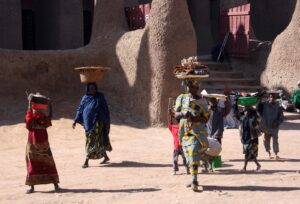 Djenne 3-Day Mosque and Market Tour from Bamako A group of people, mostly women and children, are carrying items on their heads through a dusty village with unique mud-brick buildings.