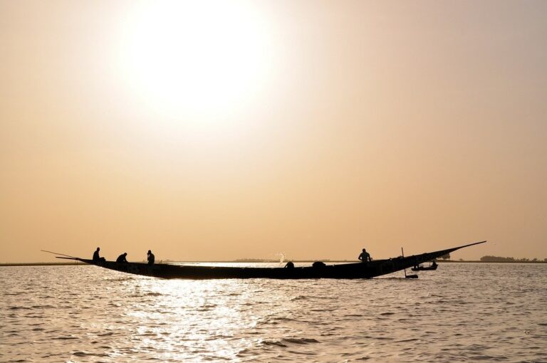 A silhouette of a long boat with several people aboard floats on calm waters under a bright, hazy sky.