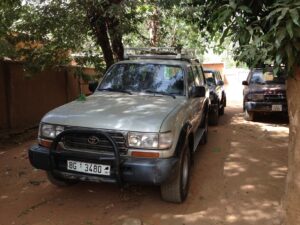 Mali 4×4 SUV Rental – Vehicle Hire Three Toyota Land Cruisers are parked on a dirt road shaded by trees. The silver one in the front has the license plate "BG 3480".