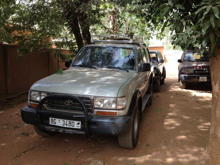 Three Toyota Land Cruisers are parked on a dirt road shaded by trees. The silver one in the front has the license plate "BG 3480".