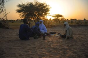 Mopti to Ancient City of Timbuktu 4-Day Trip via Niger River Four men are sitting on the sand in a desert landscape at sunset, gathered around a small pot.