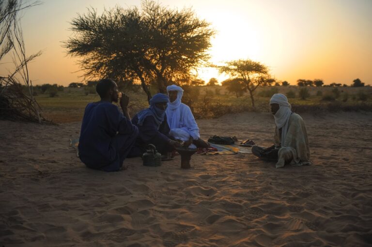 Four men are sitting on the sand in a desert landscape at sunset, gathered around a small pot.