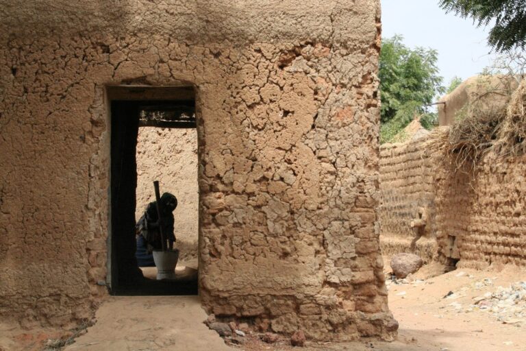 A person in dark clothing is using a mortar and pestle inside a mud-brick structure with a doorway, surrounded by textured walls and a sandy ground.