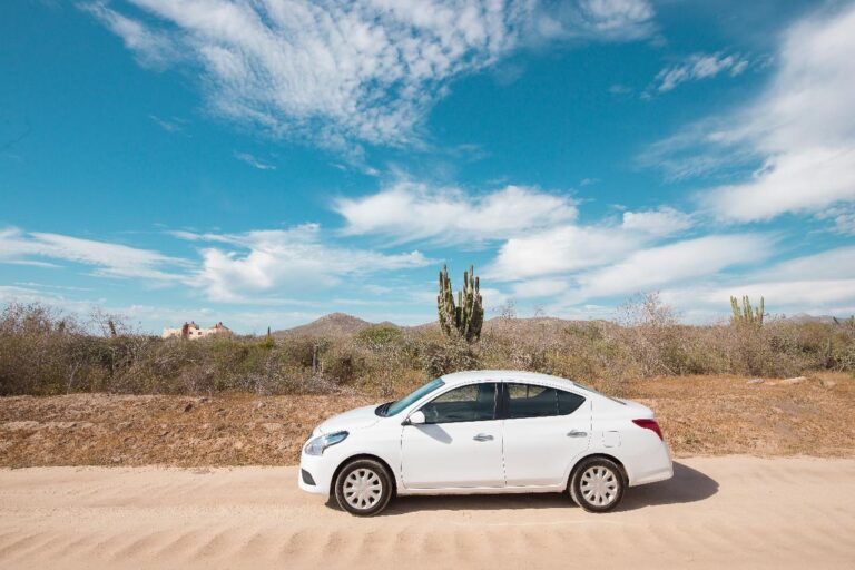 A white car is parked on a dirt road in a desert landscape with cacti, hills, and a cloudy blue sky.