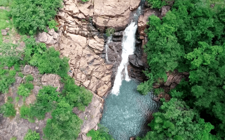 An aerial view of a waterfall cascading down a rocky cliff into a turquoise pool surrounded by lush greenery.