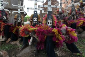 Dogon Country ‘Jewel of West Africa’ 4-Day Tour from Mopti Several people are wearing traditional Dogon masks with bright pink and yellow raffia costumes, seated on the ground outdoors.
