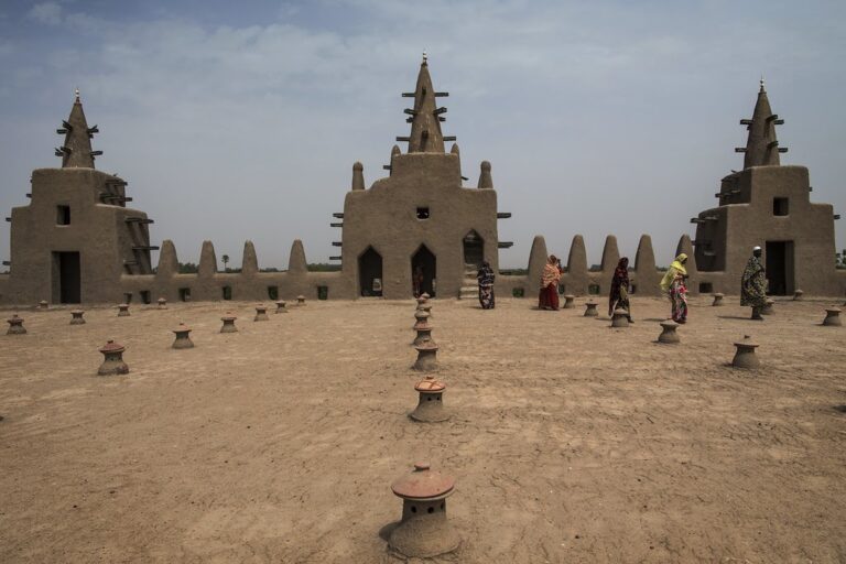 The Great Mosque of Djenné in Mali shows women in colorful dresses walking on the roof, with its iconic mud-brick architecture and distinctive conical towers.