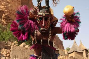 11-Day Mali Festivals Tour A person in a traditional Dogon mask and costume stands in front of a cliff dwelling, holding feathered decorations and a gourd.
